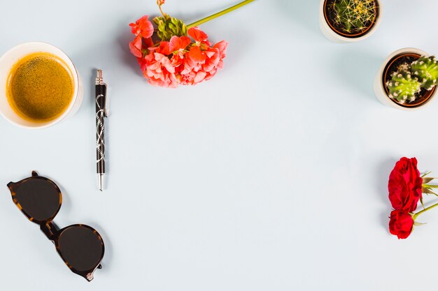 An overhead view of tea cup; pen; sunglasses; cactus potted plant and roses on white background