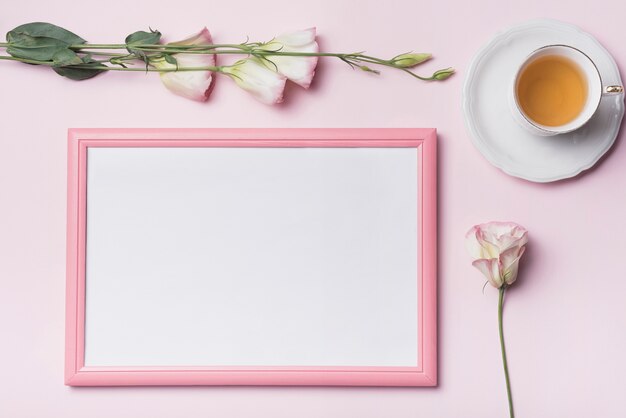 An overhead view of tea cup with picture frame and eustoma flowers against pink background