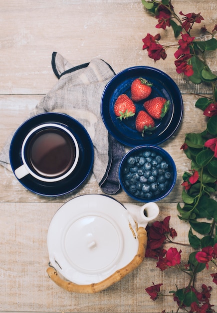 An overhead view of teapot; coffee cup; strawberries and blueberries on wooden plank