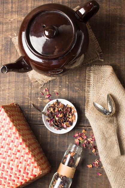 An overhead view of teapot with dried herbs; wicker box; strainer and jute on wooden desk