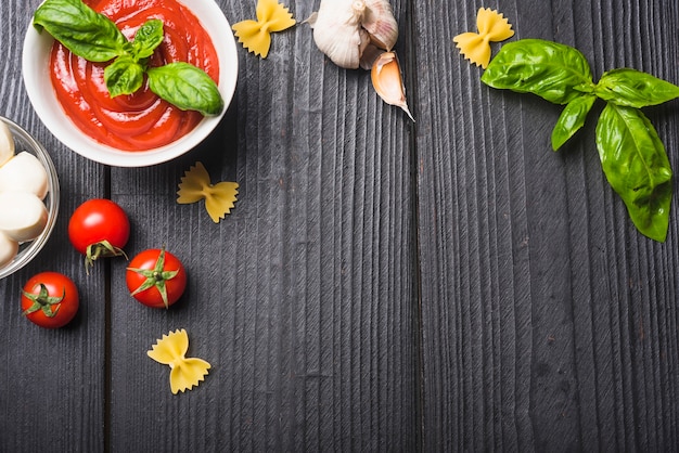 An overhead view of tomato sauce with mozzarella; pasta; garlic an basil on wooden plank