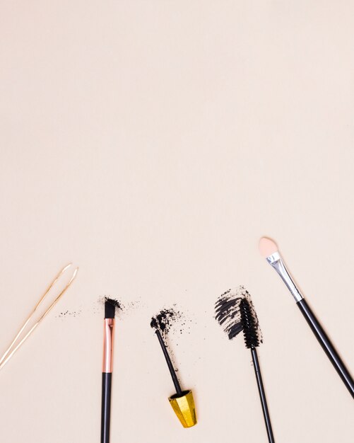 An overhead view of tweezers; makeup brush and mascara brush isolated on beige background