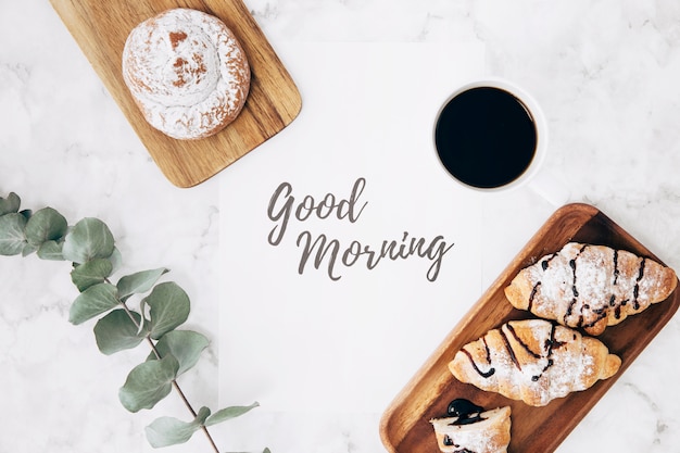 Free photo an overhead view of twig; coffee; bun and croissants with good morning message over marble texture backdrop