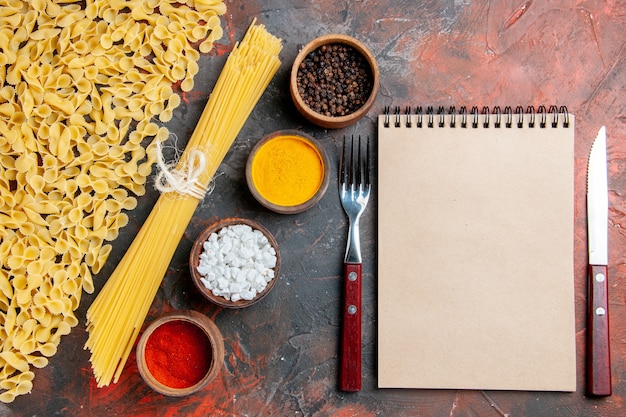 Overhead view of uncooked pasta in various forms different spices and notebook with fork and knife on black table