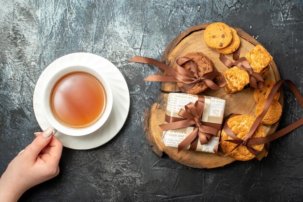 Overhead view of various delicious cookies on wooden cutting board and hand holding a cup of coffee on dark icy background