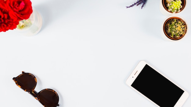 An overhead view of vase; sunglasses; cell phone and cactus potted plant on white background