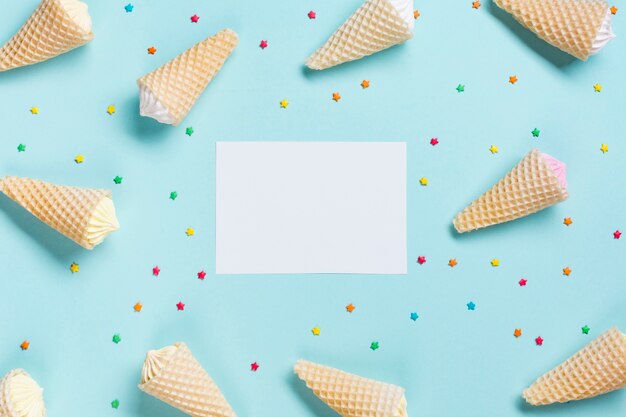 An overhead view of waffle cones and sprinkles surrounded near the white blank paper on blue backdrop