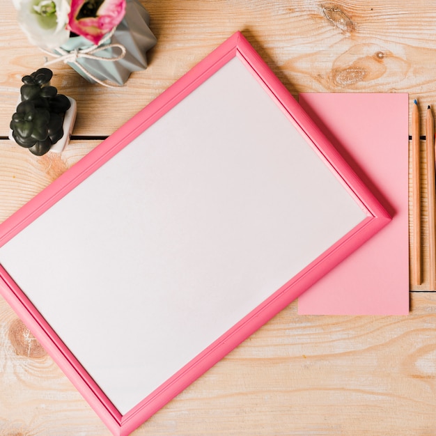 ピンクの境界線を持つ白い空白のフレームのオーバーヘッドビュー。紙;木製の机の上に色鉛筆と花瓶