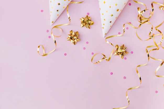 An overhead view of white paper hat; sprinkle; golden bow and streamer on pink background