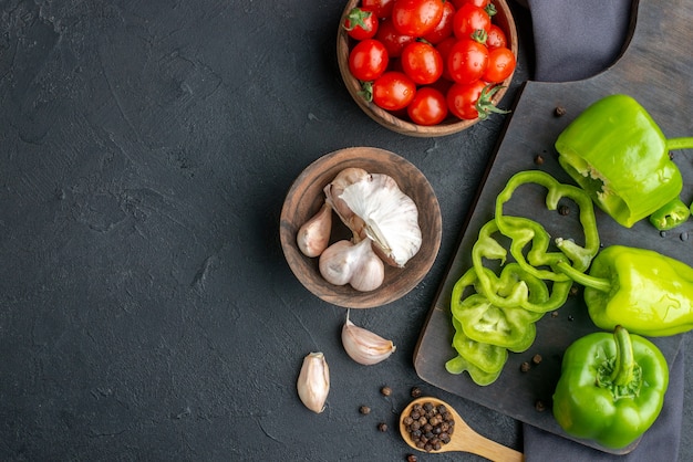 Overhead view of whole cut chopped green peppers on wooden cutting board tomatoes in bowl garlics on dark color towel on the left side on black surface
