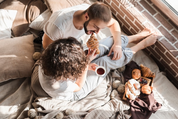 Free photo an overhead view of a woman feeding croissant to her husband sitting on bed