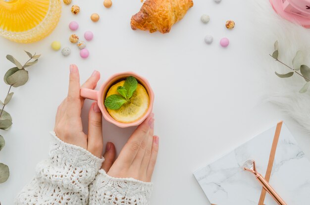 An overhead view of woman holding cup of lemon tea on white table with croissant and candies on white background