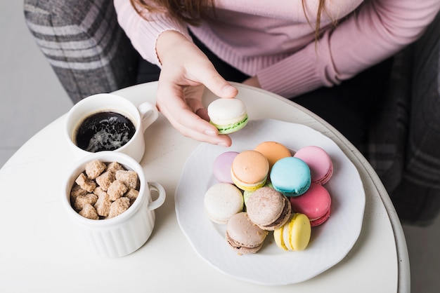 Una vista ambientale della donna che tiene maccherone con la tazza dei cubi dello zucchero bruno e caffè sulla tavola bianca