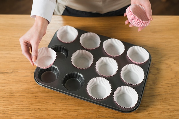 Free photo an overhead view of woman's hand arranging cupcake holder in the baking tray on wooden table