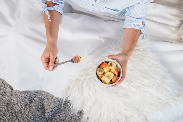 An overhead view of woman sitting in bed holding bowl of fruit salad