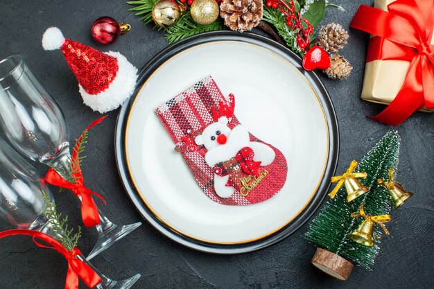 Overhead view of xsmas sock on dinner plate christmas tree fir branches conifer cone gift box santa claus hat fallen glass goblets on black background
