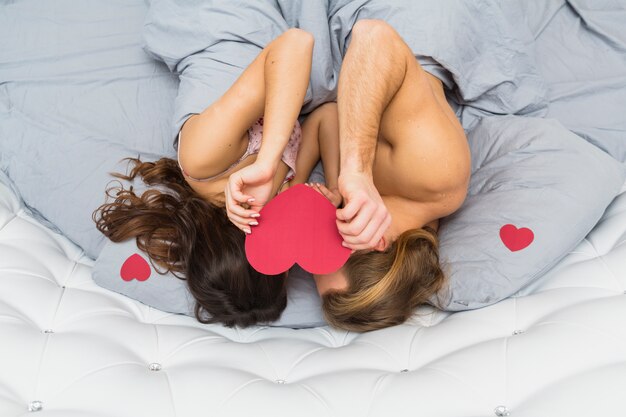 Вид сверху молодая пара спит на кровати, держа бумагу в форме сердца на их лице