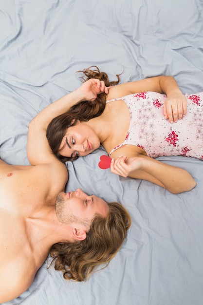 An overhead view of young woman holding heart shape paper lying near her sleeping boyfriend