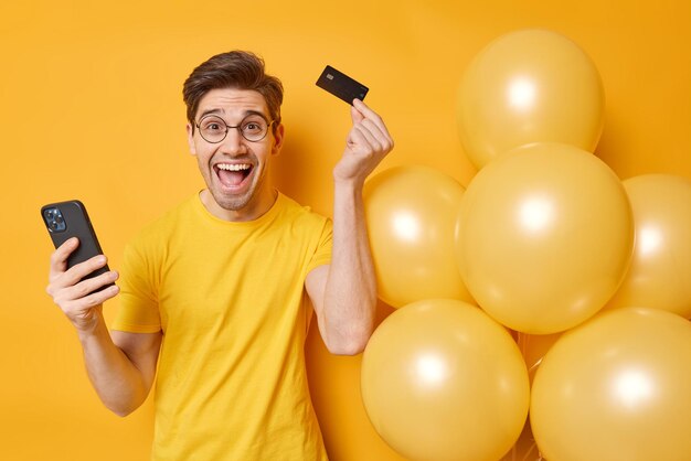Обрадованный взрослый мужчина, рад получить деньги на свой банковский счет, держит смартфон, а кредитная карта организует вечеринки рядом с кучей надутых воздушных шаров на желтом фоне. Концепция праздника