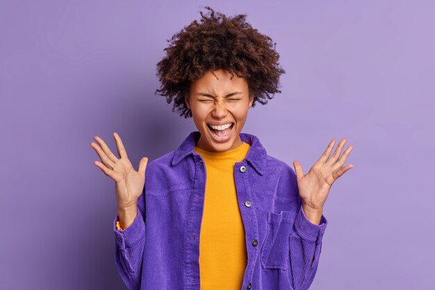 大喜びのアフリカ系アメリカ人の女の子は、ベルベットのジャケットに身を包んだ素晴らしいニュースを聞いて興奮し、とても幸せに手を上げたまま大声で叫びます。