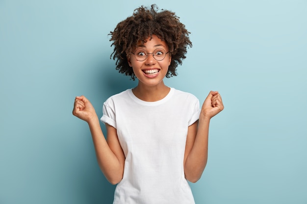 Overjoyed Afro American woman anticipates for victory, clenches fists, has broad smile