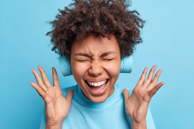 Overjoyed Afro American woman keeps hands raised exclaims joyfully closes eyes from happiness reacts on awesome news wears wireless headphones on ears isolated over blue wall. Joy concept