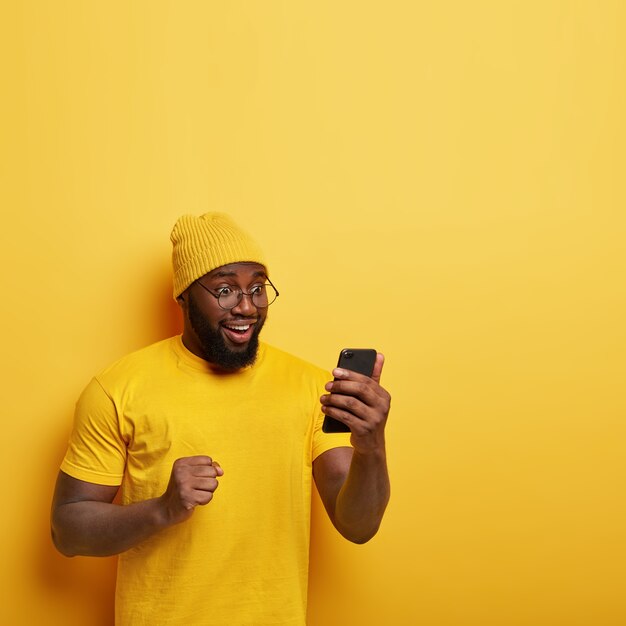 두꺼운 강모를 가진 기뻐하는 아프리카 남자, 행복하게 휴대 전화를 바라보고, 낙관적 인 느낌, 좋은 소식을 축하하고, 주먹을 움켜 쥐고, 노란색 세련된 모자를 쓰고 있습니다.
