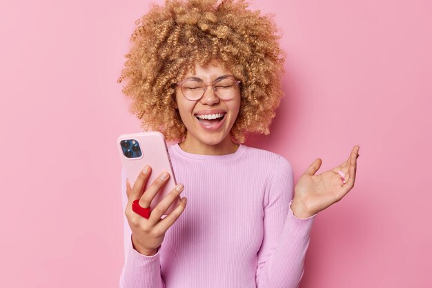 Overjoyed attractive woman with curly hair raises palm and laughs out happily wears transparent glasses and jumper watches funny video on smartphone isolated over pink background Technology