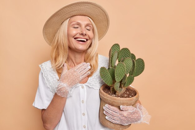 Overjoyed beautiful woman with fair hair laughs happily keeps eyes closed wears white dress lacy gloves hat carries pot of green succulent cactus isolated over beige wall