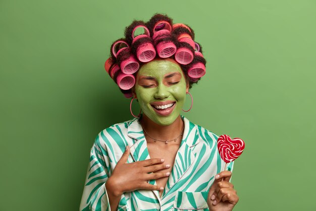Overjoyed beautiful woman with hair curlers and green facial mask, laughs happily, has fun during beauty procedures at home, holds lillopop on stick, prepares for party. Skin pampering, wellness, spa