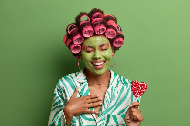 머리카락 curlers와 녹색 얼굴 마스크를 가진 기뻐하는 아름다운 여인은 행복하게 웃고 집에서 미용 절차를 즐기며 막대기에 lillopop을 들고 파티를 준비합니다. 피부 웰빙, 웰빙, 스파