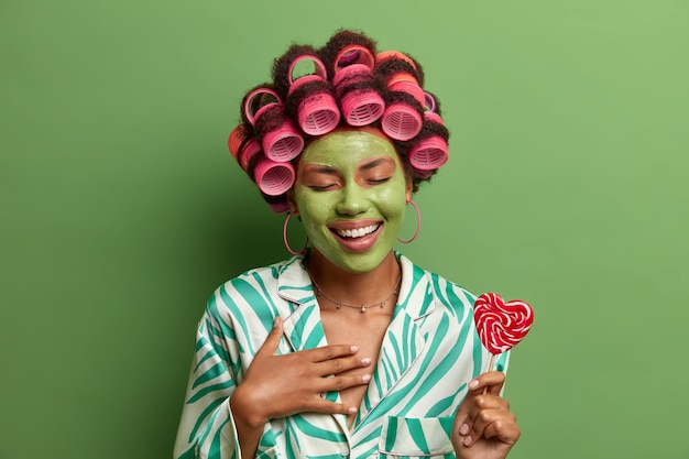 Bella donna felicissima con bigodini e maschera facciale verde, ride allegramente, si diverte durante le procedure di bellezza a casa, tiene il lillopop sul bastone, si prepara per la festa. coccole della pelle, benessere, spa