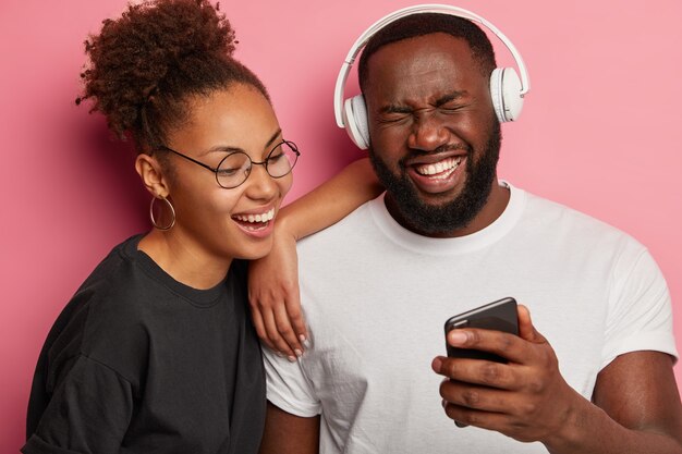 Overjoyed black hipster guy holds smartphone, laughs happily as watch funny movie on smartphone together with girlfriend, use stereo headphones.