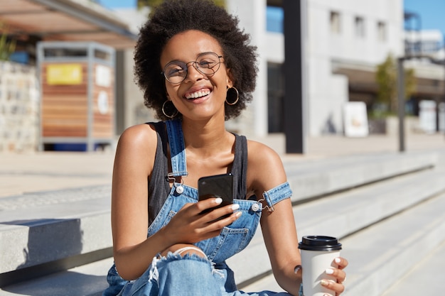 大喜びの黒人女性は面白い表情で笑い、スマートフォンのソーシャルネットワークで逸話を読み、持ち帰り用のコーヒーを飲み、スタイリッシュな服を着ています。混血の女性が国際電話を待つ