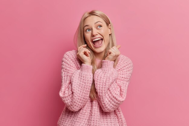 Overjoyed blonde young woman has happy face expression looks above keeps mouth opened dressed in casual clothes