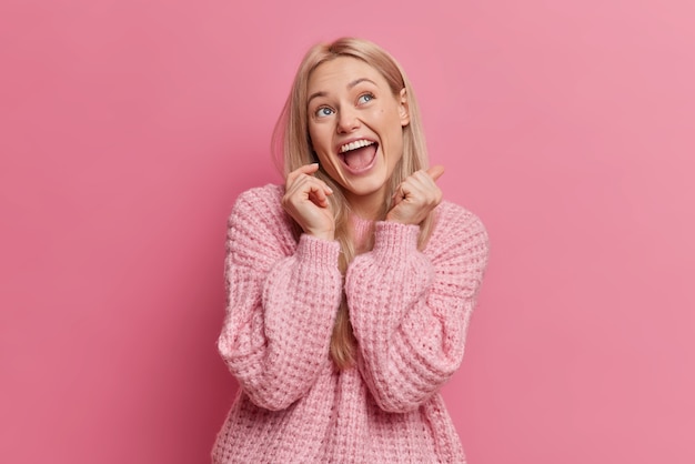 Overjoyed blonde young woman has happy face expression looks above keeps mouth opened dressed in casual clothes