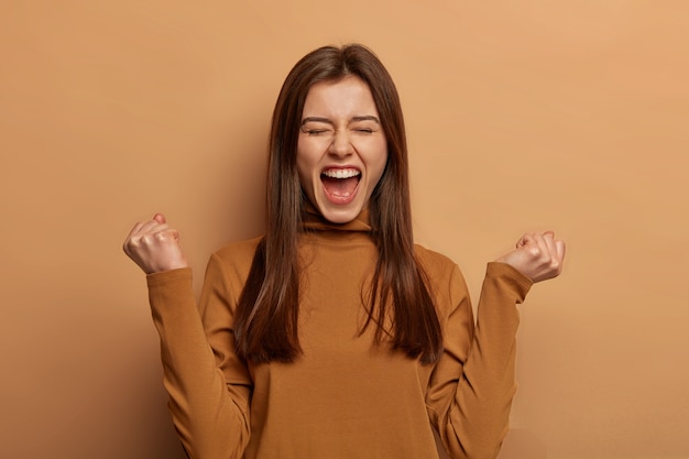 Overjoyed brunette woman make fists pump, celebrates success, shouts and feels like champion, keeps mouth wide opened, wears turtleneck