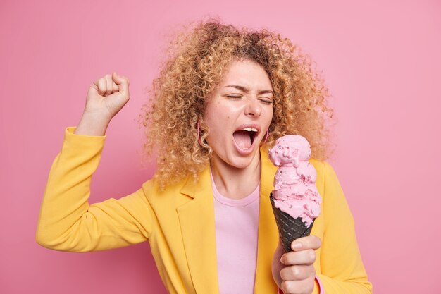Overjoyed carefree curly haired woman has fun sings and dances with tasty ice cream of strawberry flavor raises clenches fist dressed in yellow jacket has sweet tooth. Junk food summer time concept
