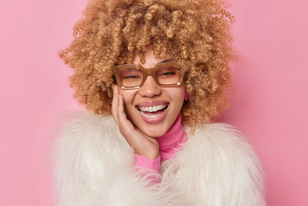 Overjoyed curly haired woman giggles positively keeps hand on cheek laughs out feels very positive wears transparent glasses and winter coat expresses positive emotions isolated on pink wall