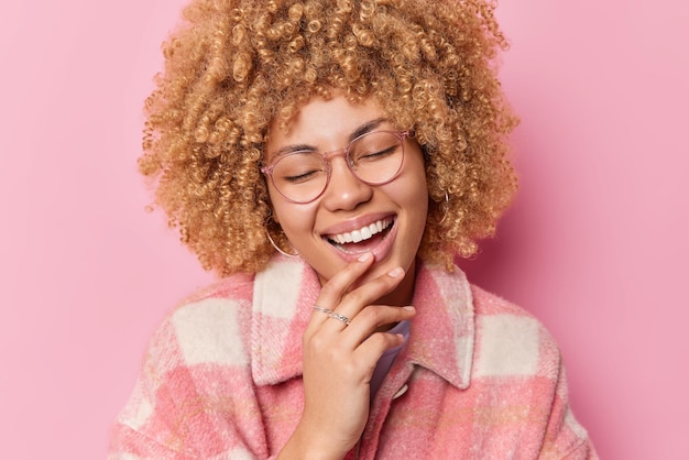 大喜びの縮れ毛の女性は積極的に笑う笑顔は広くあごに手を保ちます良い気分である透明な眼鏡とピンクの背景の上に分離されたスタイリッシュなジャケットを着ています幸せな感情