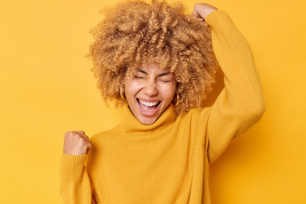 大喜びの巻き毛の女性は、はいジェスチャーを興奮させ、喜びから勝利を叫びます黄色のスタジオの背景の上にカジュアルなセーターのポーズを着て成功を祝いますはい私は私の目標を達成しました