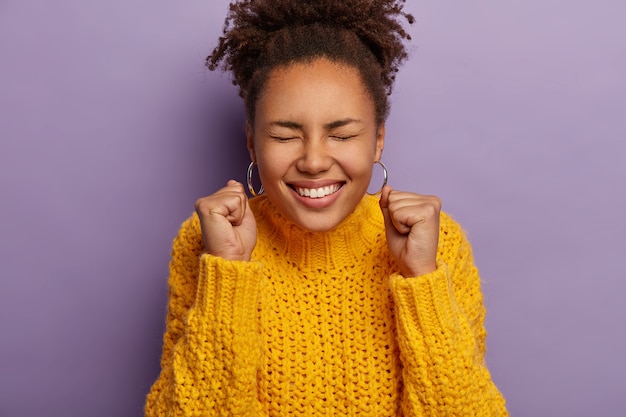 Free photo overjoyed curly haired woman raises clenched fists, feels excited, celebrates success, wears warm yellow knitted sweater