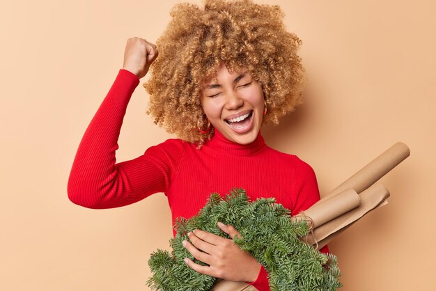 Overjoyed curly woman celebrates good news makes fist bump and exclaims happily holds green spurce wreath and paper for wrapping presents wears red turtleneck isolated over beige background.