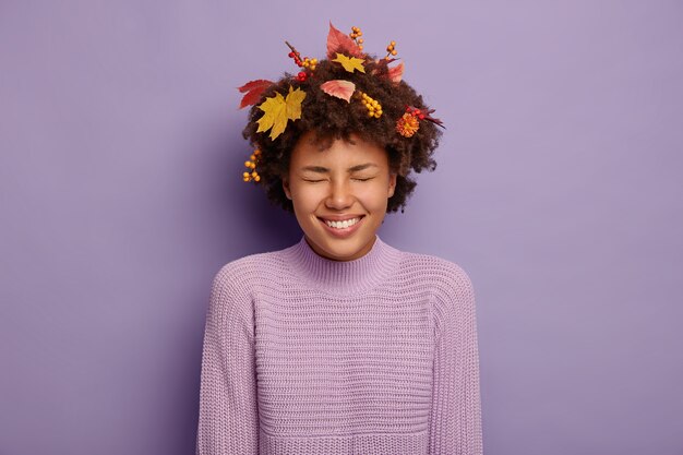 Overjoyed curly woman laughs positively, has autumn mood, smiles broadly, jokes around with friends, has yellow maple leaves and rowan berries in hair, dressed in casual wear. Happiness, wellbein
