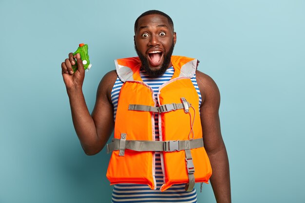 Overjoyed dark skinned man has water fighting with friends at beach, exclaims happily, wears orange lifejacket