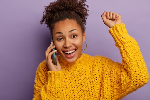 Overjoyed dark skinned woman has positive mobile conversation, hears excellent news, raises arm and dances from joy