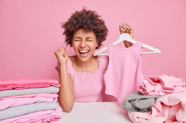 巻き毛の黒い肌の女性が幸せから拳を握りしめ、ハンガーにアパレルを保持し、ピンクの壁に隔離された洗濯物をテーブルのひだに座っている