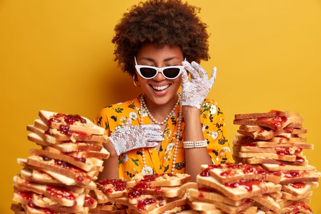 Overjoyed elegant lady with Afro hair smiles happily wears stylish sunglasses poses against pile of tasty sandwiches