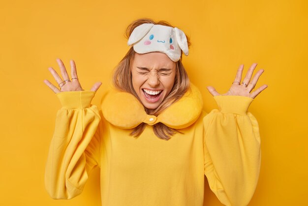 Overjoyed emotional woman raises palms exclaims loudly from joy keeps eyes closed wears sleepmask neck pillow reacts on something awesome isolated over bright yellow background. Reaction concept