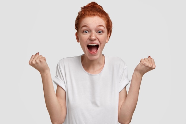 Free photo overjoyed emotive freckled woman with ginger hair, raises fists and exclaims loudly, cheers up friend, shouts supportive words, wears casual mockup white t shirt, models and gestures